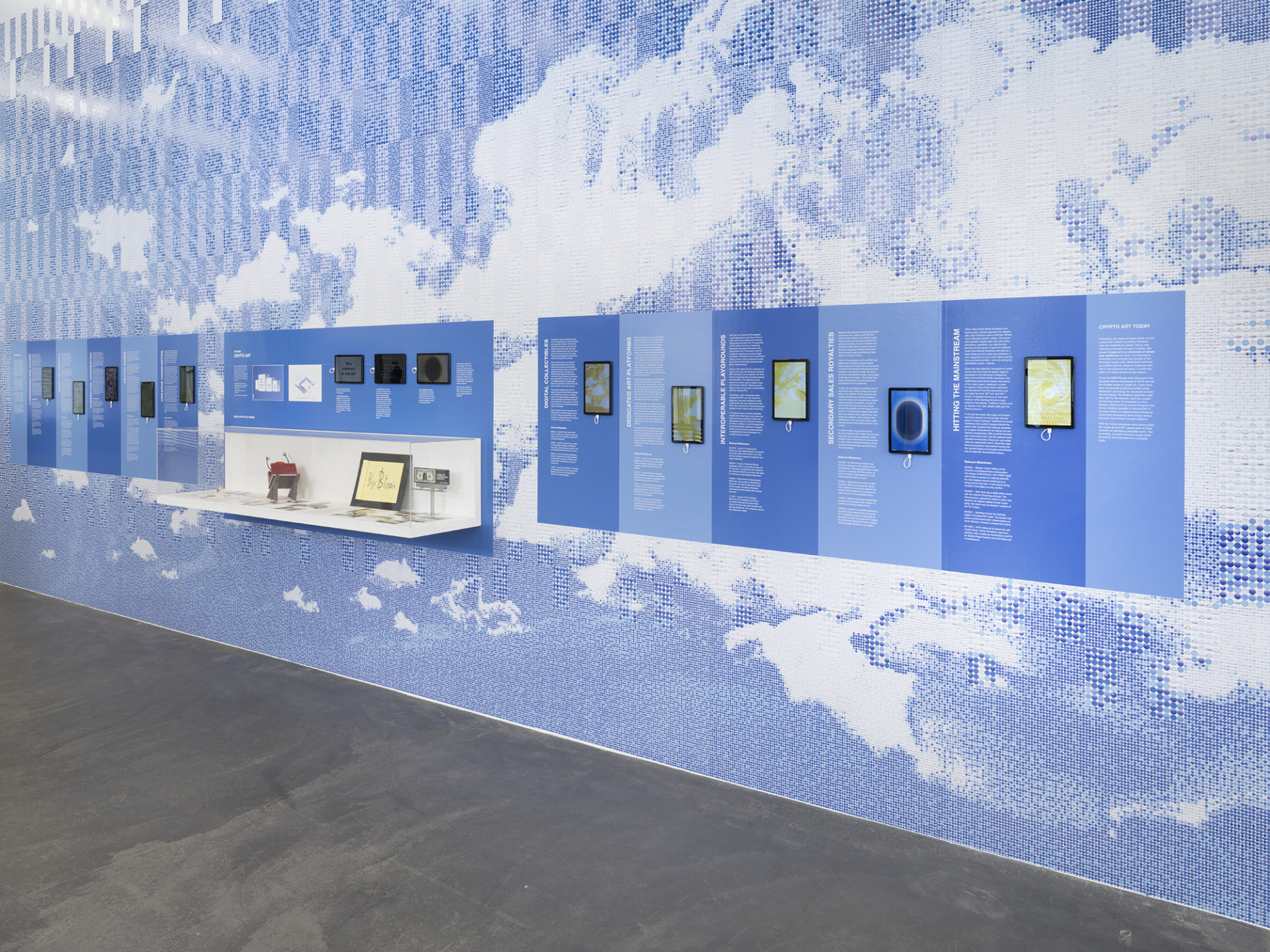 A view of an installation on a wall featuring several text-heavy printed panels, with blue and white designs evoking clouds with ASCII art