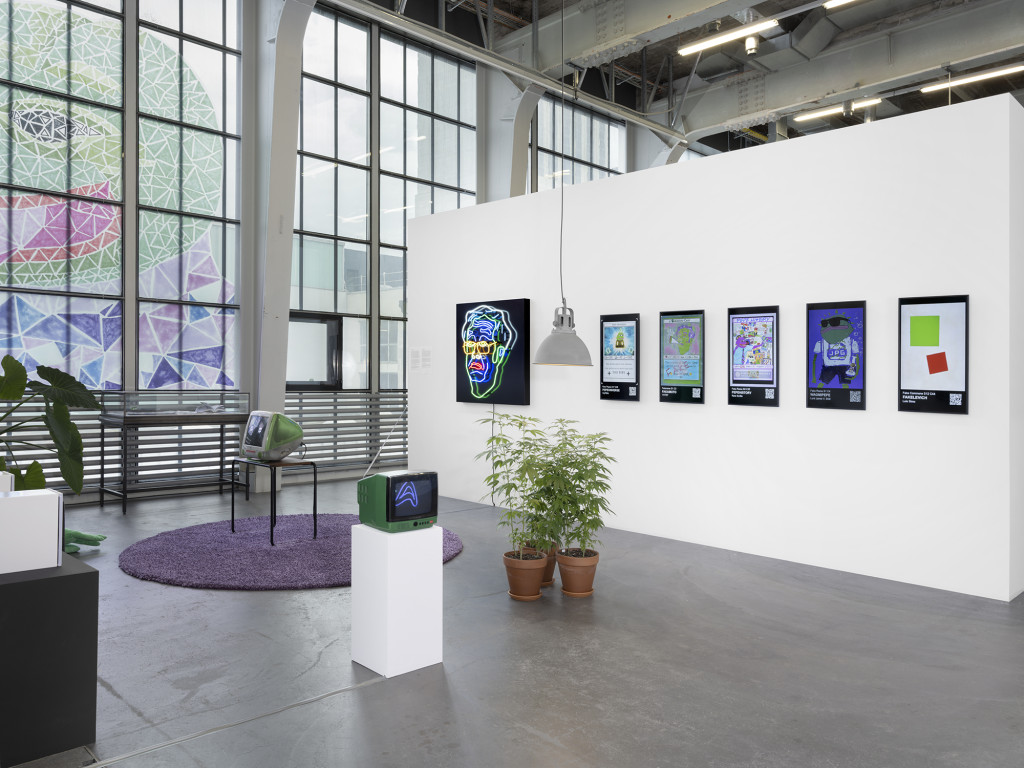 A view of an installation in a gallery featuing prints and a neon sclupture on a wall and two obsoloete monitors on plints, with a rug and houseplants creating a domestic atmosphere
