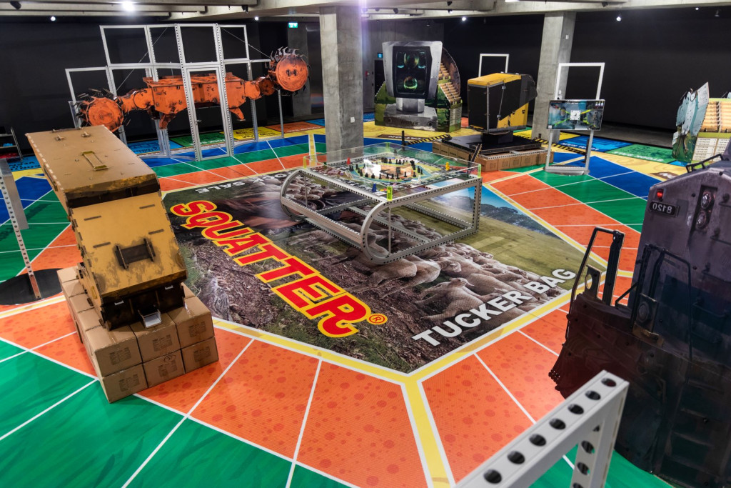 A photograph of an exhibition featuring large sculptures of cardboard and metal representing mining equipment and other technological devices, arranged on a floor designed to look like a game board