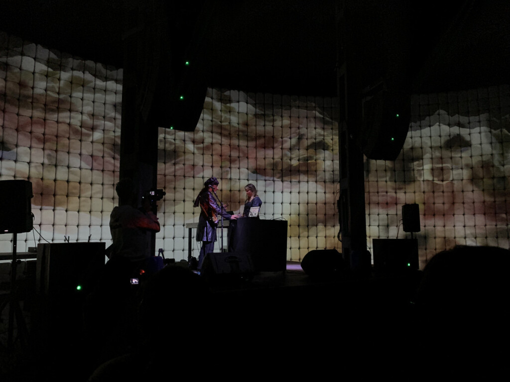 A photograph of a live performance, showing two women standing at a podium against a backdrop of projected abstract images