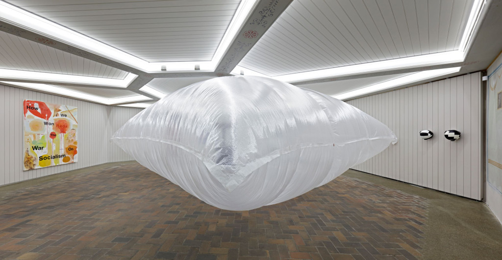 A large white inflatable cushion fills the middle of a white room; artworks hang on the walls to either side
