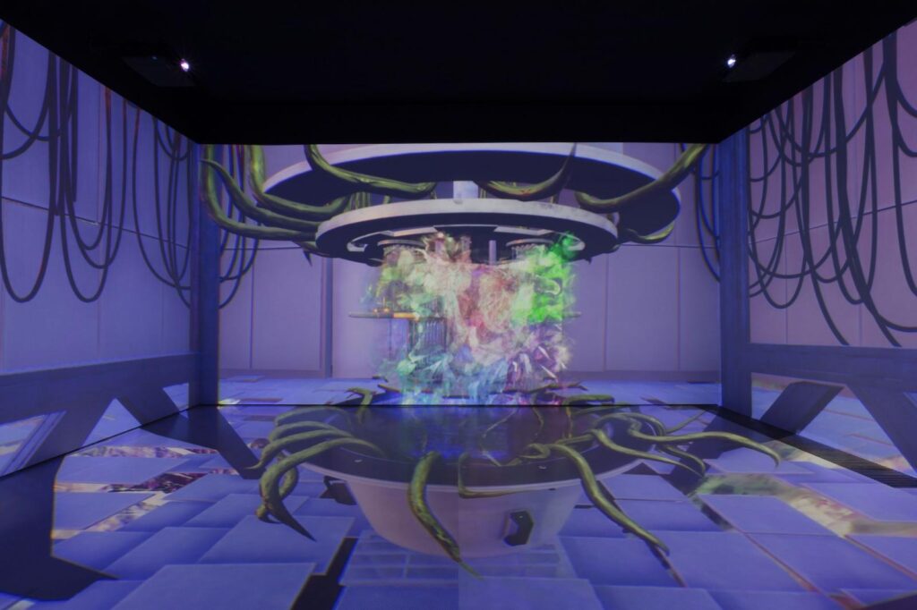 A photograph of an installation in which a purple futuristic video is projected onto three walls and the floor