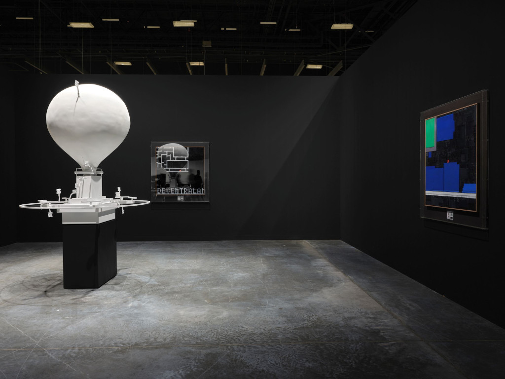 A photograph of several artworks under lights in a dark room. At the center is a sculpture of a drone balloon, and abstract geometric paintings hang on the black walls