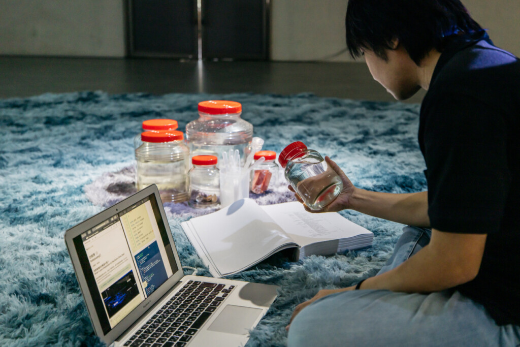 A photo of a person sitting on a blue rug in front of a Macbook, on which are laid red-lidded jars of water