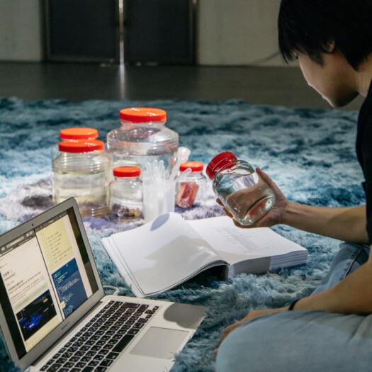 A photo of a person sitting on a blue rug in front of a Macbook, on which are laid red-lidded jars of water