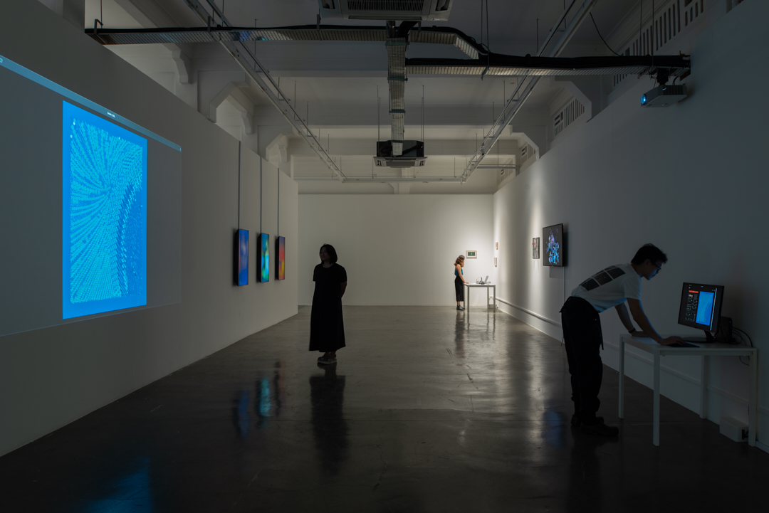 A view of a gallery with white walls, with a mix of projections, flatscreens and computer screens showing artworks, and people milling about or interacting with works