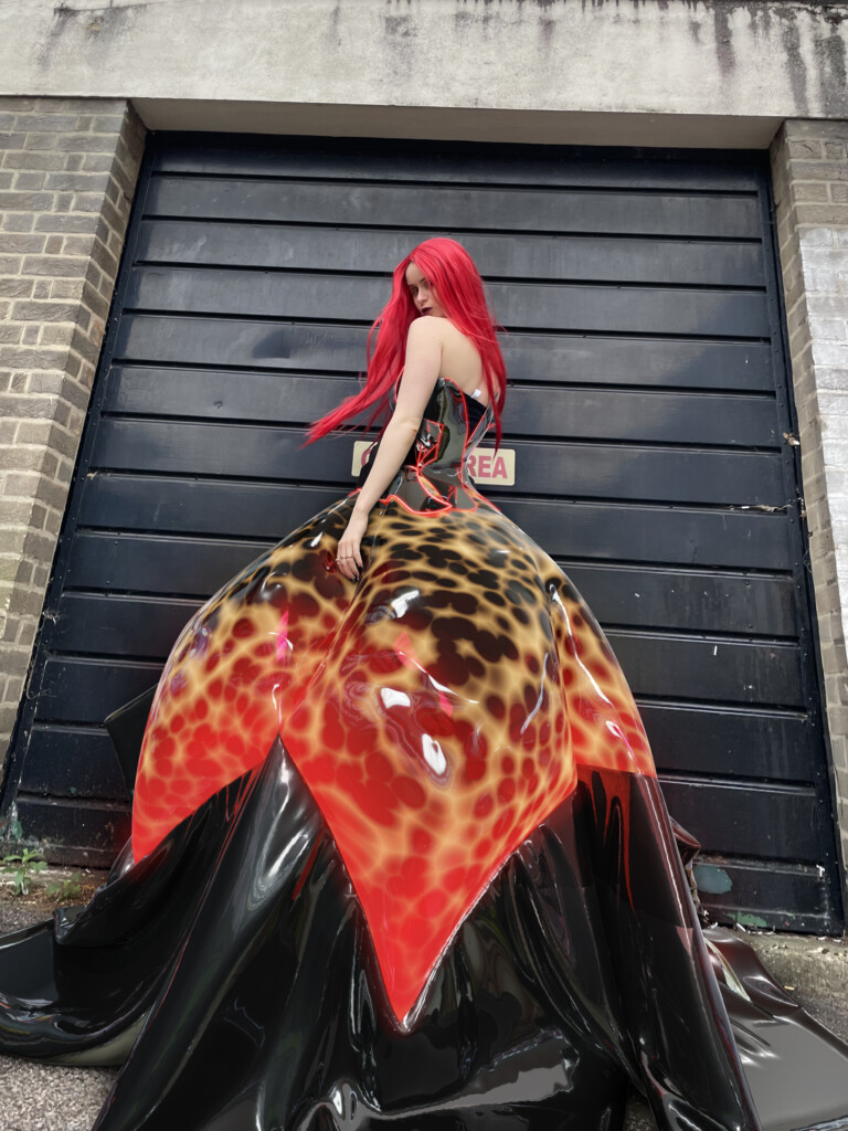 A pale woman in a red wig poses in a flamboyant digital dress with leopard print and shiny black hem against a garage door