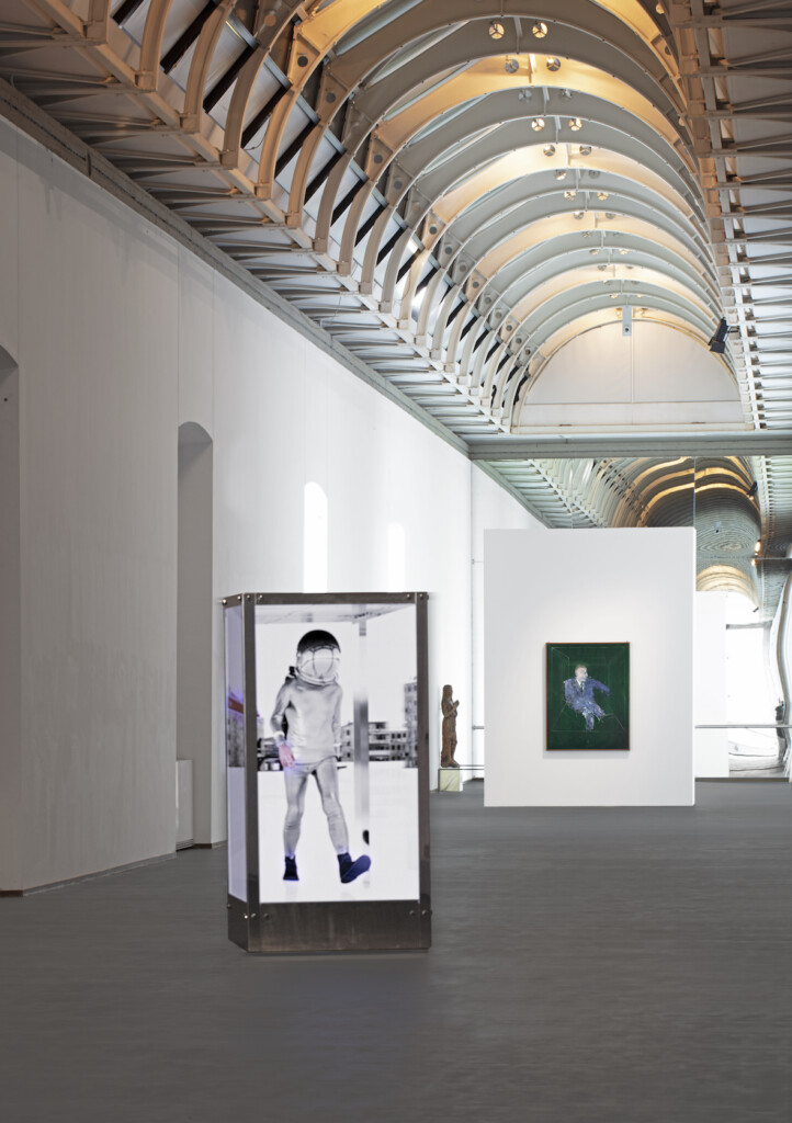 A metallic booth displaying a digital image of a walking astronaut stands in a long gallery with a vaulted ceiling