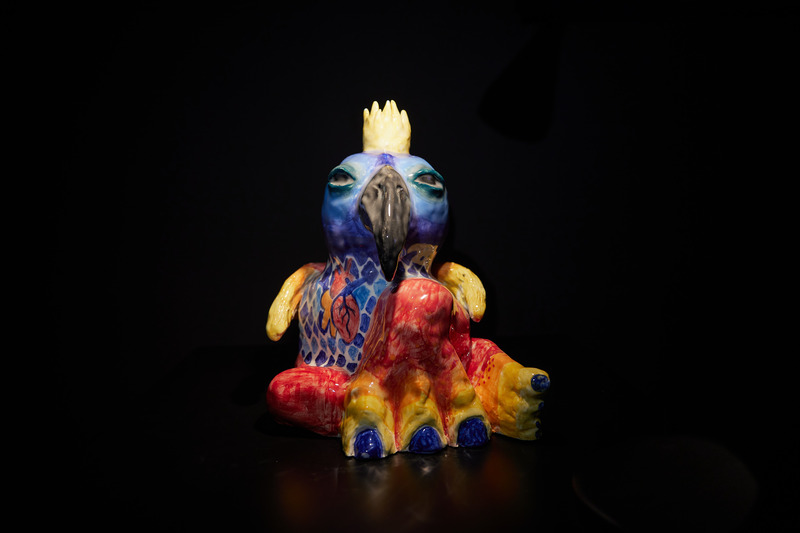 A ceramic sculpture of a bird with a black beak and blue and red plumage against a black background.