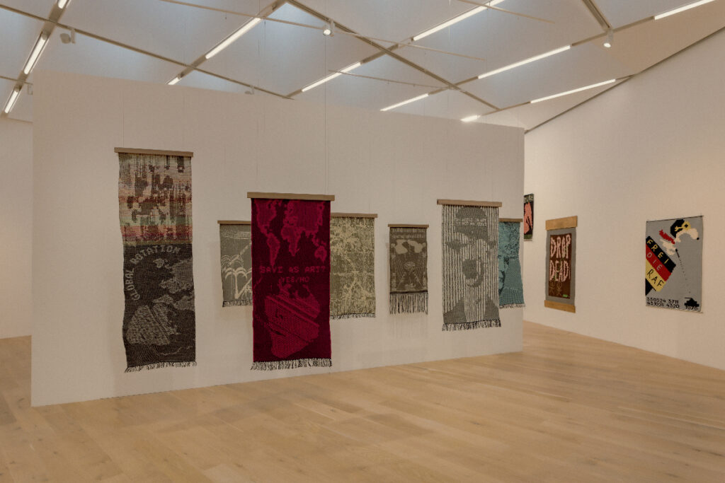 A photo of a gallery with white walls and wooden floors, in which a series of vertical tapestries hang from the ceiling