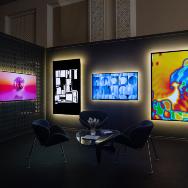 A booth at an art fair with backlit monitors on black walls showing colorful and striking abstract digital art.