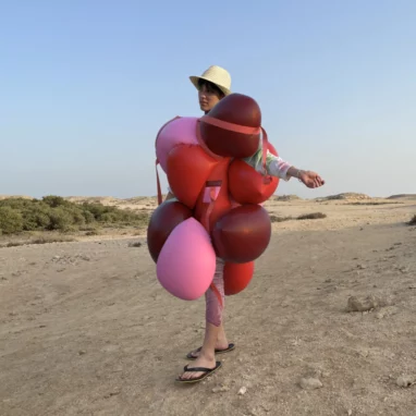 A person stands with arms outstretched in an arid desert landscape, appearing to wear a dress made from red and pink balloons