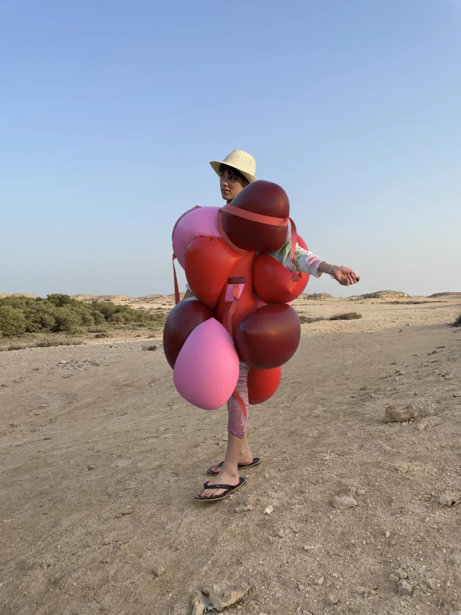 A person stands with arms outstretched in an arid desert landscape, appearing to wear a dress made from red and pink balloons