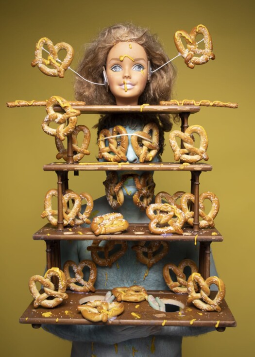 A surreal photograph of a man painted in blue paint with a shelving unit covered in pretzels and crowned with a doll's head attached to him
